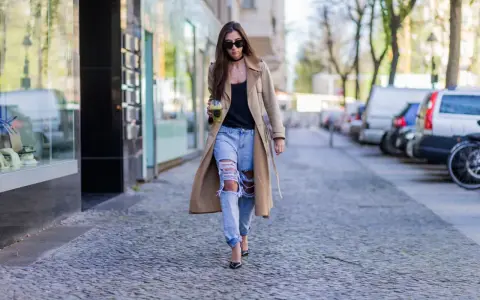 Frau in Trenchcoat und zerrissenen Boyfriend Jeans kombiniert mit Pumps und Sonnenbrille geht mit Getränk.