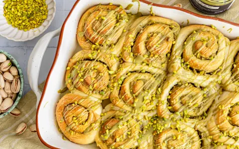 Frisch gebackene Pistazienschnecken, beträufelt mit Pistaziencreme und bestreut mit gehackten Pistazien. Daneben Schalen mit ganzen und gehackten Pistazien.