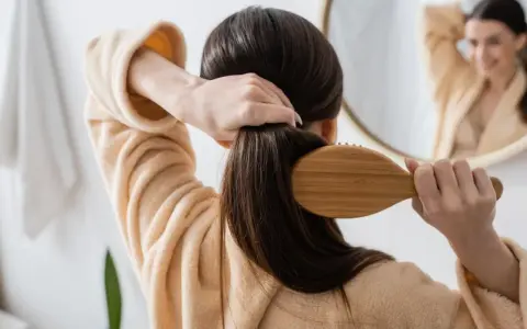 Frau kämmt ihr langes, dunkles Haar mit einer Holzbürste. Sie trägt einen flauschigen Bademantel. Tipps, wie wachsen haare schneller, sind hier nicht sichtbar.