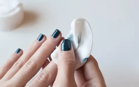 Eine Hand mit dunkelblauem Nagellack wird mit einem Wattepad abgewischt, um den Nagellack zu entfernen.