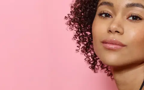 Ein Porträt einer jungen Frau mit vollen, glänzenden Lippen und lockigem Haar vor einem rosafarbenen Hintergrund. Ein Hauch von Make-up, das 50 Jahre Erfahrung widerspiegelt.