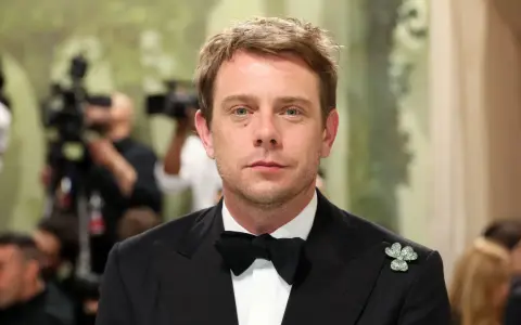Jonathan Anderson trägt einen schwarzen Smoking mit Fliege und einem Kleeblatt-Anstecker. Im Hintergrund sind Kameras und unscharfe Personen zu sehen.