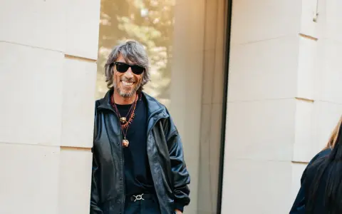 Ein Mann mit grauen Haaren und Sonnenbrille, gekleidet in eine Lederjacke, lächelt. Er könnte ein Balenciaga Designer sein, der seinen Stil zur Schau stellt.