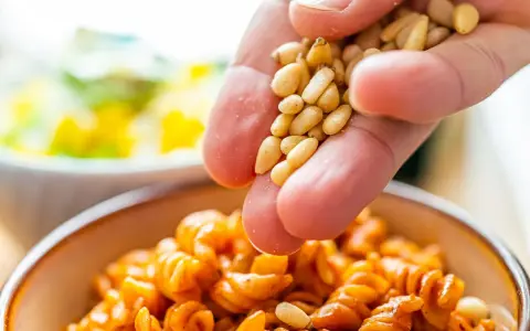 Eine Hand streut Pinienkerne über eine Schüssel mit Pasta. Pinienkerne gesund und lecker!