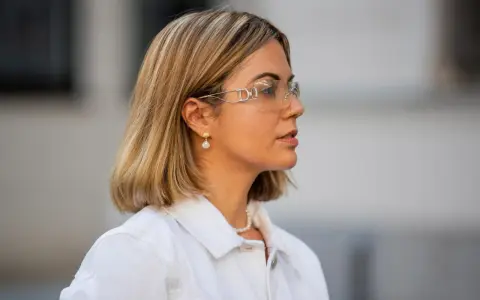 Eine Frau mit einem schicken Bob und auffälligen Dior-Sonnenbrillen. Sie trägt eine weiße Jeansjacke und Perlenschmuck.