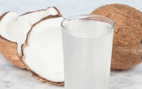 Ein Glas erfrischendes Kokoswasser neben einer ganzen und einer halbierten Kokosnuss. Dieses natürliche Getränk ist bekannt dafür, dass Kokoswasser gesund ist.