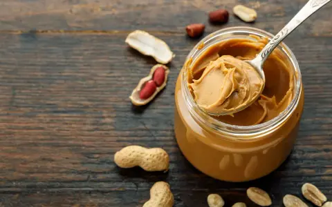 Ein Löffel cremiger Erdnussbutter ragt aus einem Glas. Umgeben von Erdnüssen und Schalen, zeigt das Bild, wie erdnussbutter gesund sein kann.