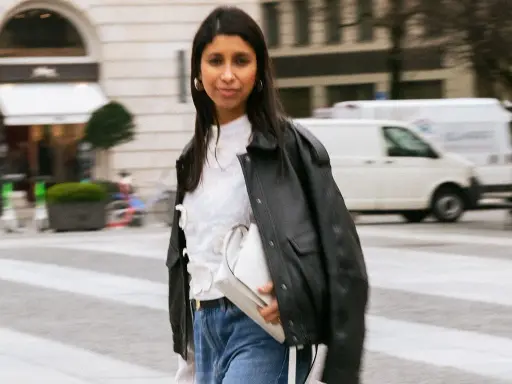 Stylische Frau kombiniert eine weiße Bluse mit Jeans und Lederjacke. Sie trägt eine weiße Tasche und goldene Ohrringe.