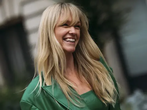 Lachende Frau mit blonden, langen Haaren und einem leichten Pony, trägt ein grünes Outfit.