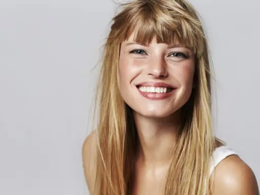 Strahlendes Lächeln einer jungen Frau mit einer lässigen Pony Frisur und langen blonden Haaren.