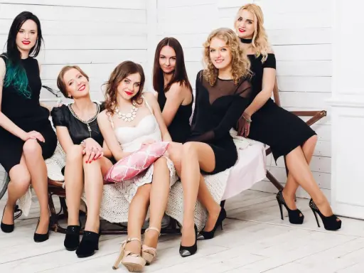 Sechs Frauen in eleganten Kleidern, die meisten davon schwarz, sitzen auf einer Bank. Eine Brautjungfer in Weiß hält ein rosa Kissen. Ein stilvolles Bild für eine Hochzeit.