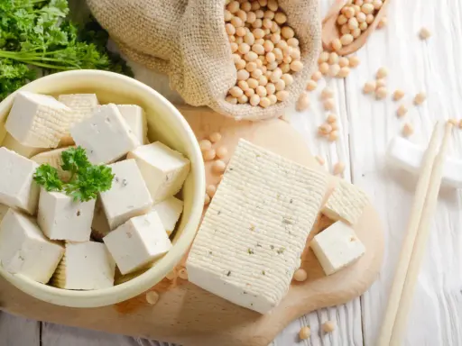 Würfel und ein Block Tofu liegen auf einem Holzbrett. Sojabohnen in einem Sack und Petersilie deuten auf die tofu inhaltsstoffe hin.