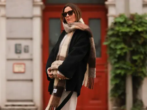 Frau mit Sonnenbrille, die einen karierten Schal lässig um den Hals gebunden hat. Sie hält eine gesteppte Tasche.
