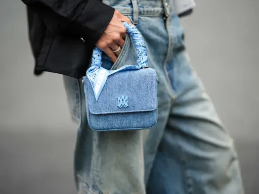 Ein hellblaues Jeans-Täschchen mit einem quadratischen Tuch binden, das den Griff ziert. Das Tuch hat ein Paisleymuster und den Schriftzug "AMIRI".