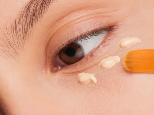 Nahaufnahme eines Auges, auf das mit einem Pinsel Concealer aufgetragen wird. Der Concealer setzt sich ab und bildet kleine Tupfen.