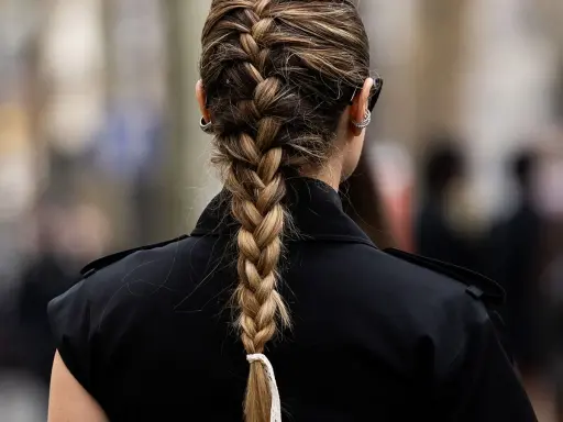 Eine Frau mit einem kunstvollen french braid, das elegant über ihre Schulter fällt. Sie trägt einen schwarzen Blazer und auffällige Ohrringe.