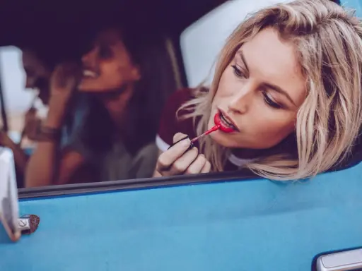 Blonde Frau mit rotem Lippenstift im Auto, um ihr Make-up aufzufrischen. Freundinnen im Hintergrund.