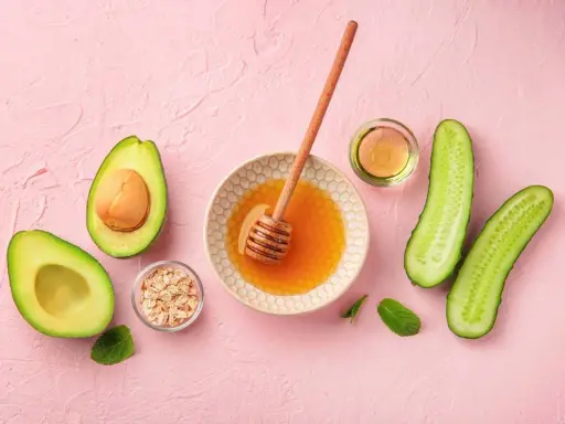 Zutaten für eine Gesichtsmaske selber machen: Avocado, Honig, Haferflocken, Gurke und Öl auf rosa Untergrund.