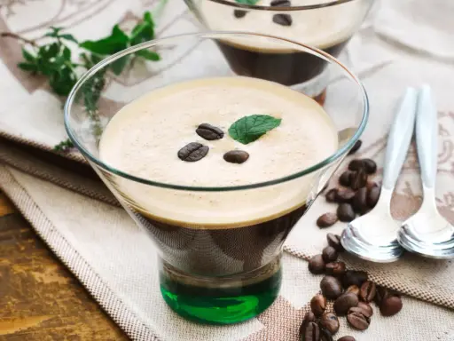 Ein Glas italienischer Eiskaffee mit Kaffeebohnen und Minze garniert, daneben Löffel und Kaffeebohnen auf einem Tuch.