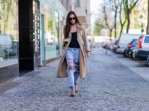 Frau in Trenchcoat und zerrissenen Boyfriend Jeans kombiniert mit Pumps und Sonnenbrille geht mit Getränk.