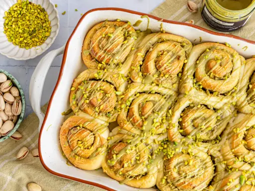 Frisch gebackene Pistazienschnecken, beträufelt mit Pistaziencreme und bestreut mit gehackten Pistazien. Daneben Schalen mit ganzen und gehackten Pistazien.