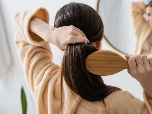 Frau kämmt ihr langes, dunkles Haar mit einer Holzbürste. Sie trägt einen flauschigen Bademantel. Tipps, wie wachsen haare schneller, sind hier nicht sichtbar.