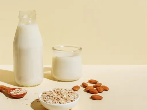 Flasche und Glas mit milchigem Getränk, daneben Mandeln und Haferflocken. Ob Mandelmilch oder Hafermilch, die Wahl ist leicht.