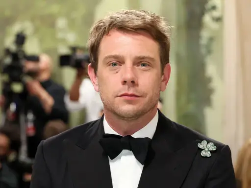 Jonathan Anderson trägt einen schwarzen Smoking mit Fliege und einem Kleeblatt-Anstecker. Im Hintergrund sind Kameras und unscharfe Personen zu sehen.