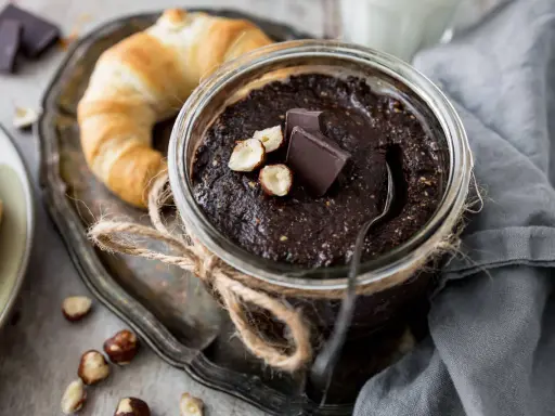 Selbstgemachte Haselnusscreme im Glas, garniert mit Schokostückchen und Nüssen, neben einem Croissant. Perfekt zum Haselnusscreme selber machen!
