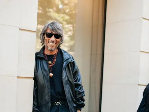 Ein Mann mit grauen Haaren und Sonnenbrille, gekleidet in eine Lederjacke, lächelt. Er könnte ein Balenciaga Designer sein, der seinen Stil zur Schau stellt.