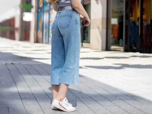 Styling culotte jeans: Ein lässiger Look mit hellblauen Jeans, weißem Shirt und Sneakern auf gepflastertem Weg.