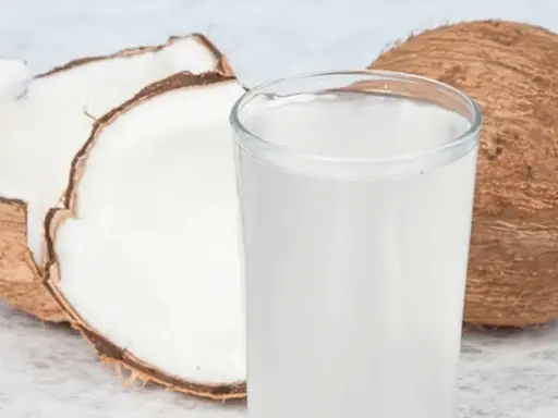Ein Glas erfrischendes Kokoswasser neben einer ganzen und einer halbierten Kokosnuss. Dieses natürliche Getränk ist bekannt dafür, dass Kokoswasser gesund ist.