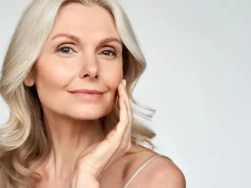 Eine Frau mit blonden Haaren berührt sanft ihr Gesicht. Ihr Make-up setzt sich in den feinen Falten ab, was ihrer Haut Tiefe verleiht.