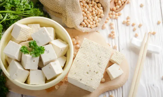 Würfel und ein Block Tofu liegen auf einem Holzbrett. Sojabohnen in einem Sack und Petersilie deuten auf die tofu inhaltsstoffe hin.
