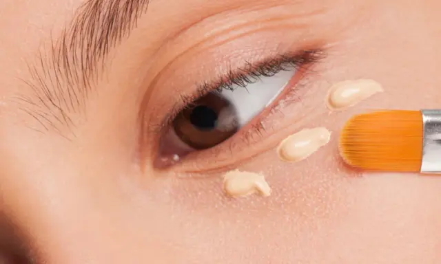 Nahaufnahme eines Auges, auf das mit einem Pinsel Concealer aufgetragen wird. Der Concealer setzt sich ab und bildet kleine Tupfen.