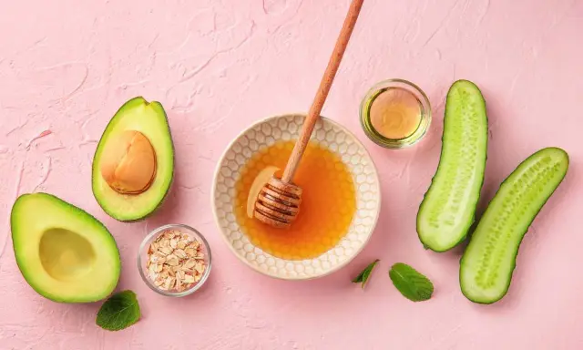 Zutaten für eine Gesichtsmaske selber machen: Avocado, Honig, Haferflocken, Gurke und Öl auf rosa Untergrund.