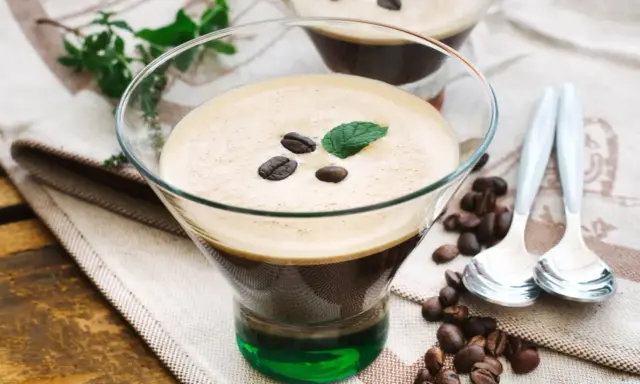 Ein Glas italienischer Eiskaffee mit Kaffeebohnen und Minze garniert, daneben Löffel und Kaffeebohnen auf einem Tuch.
