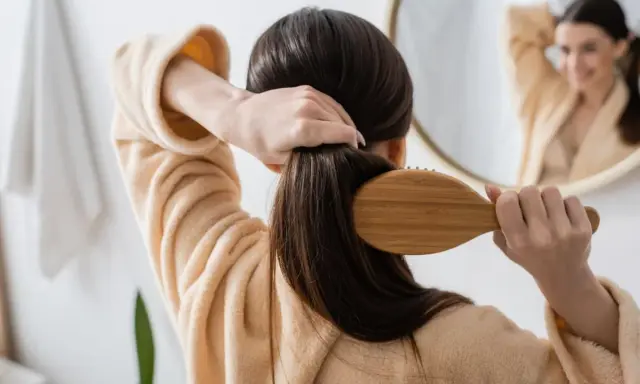 Frau kämmt ihr langes, dunkles Haar mit einer Holzbürste. Sie trägt einen flauschigen Bademantel. Tipps, wie wachsen haare schneller, sind hier nicht sichtbar.