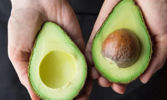 Hände halten zwei Avocadehälften. Eine Avocado mit Kern, die andere leer. Manche sagen, Avocado sei krebserregend, aber das stimmt nicht.
