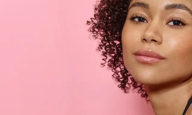 Ein Porträt einer jungen Frau mit vollen, glänzenden Lippen und lockigem Haar vor einem rosafarbenen Hintergrund. Ein Hauch von Make-up, das 50 Jahre Erfahrung widerspiegelt.