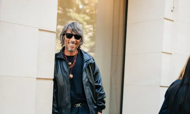 Ein Mann mit grauen Haaren und Sonnenbrille, gekleidet in eine Lederjacke, lächelt. Er könnte ein Balenciaga Designer sein, der seinen Stil zur Schau stellt.