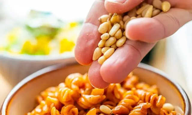 Eine Hand streut Pinienkerne über eine Schüssel mit Pasta. Pinienkerne gesund und lecker!