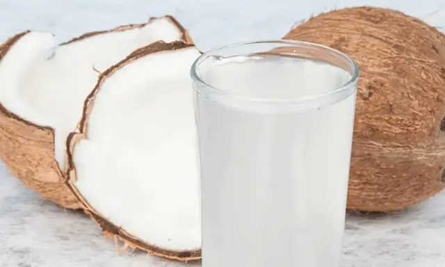 Ein Glas erfrischendes Kokoswasser neben einer ganzen und einer halbierten Kokosnuss. Dieses natürliche Getränk ist bekannt dafür, dass Kokoswasser gesund ist.