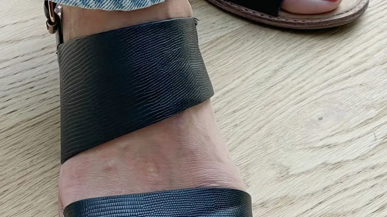Fuß in einem schwarzen Riemchensandalen mit Reptilprägung auf hellem Holzboden.