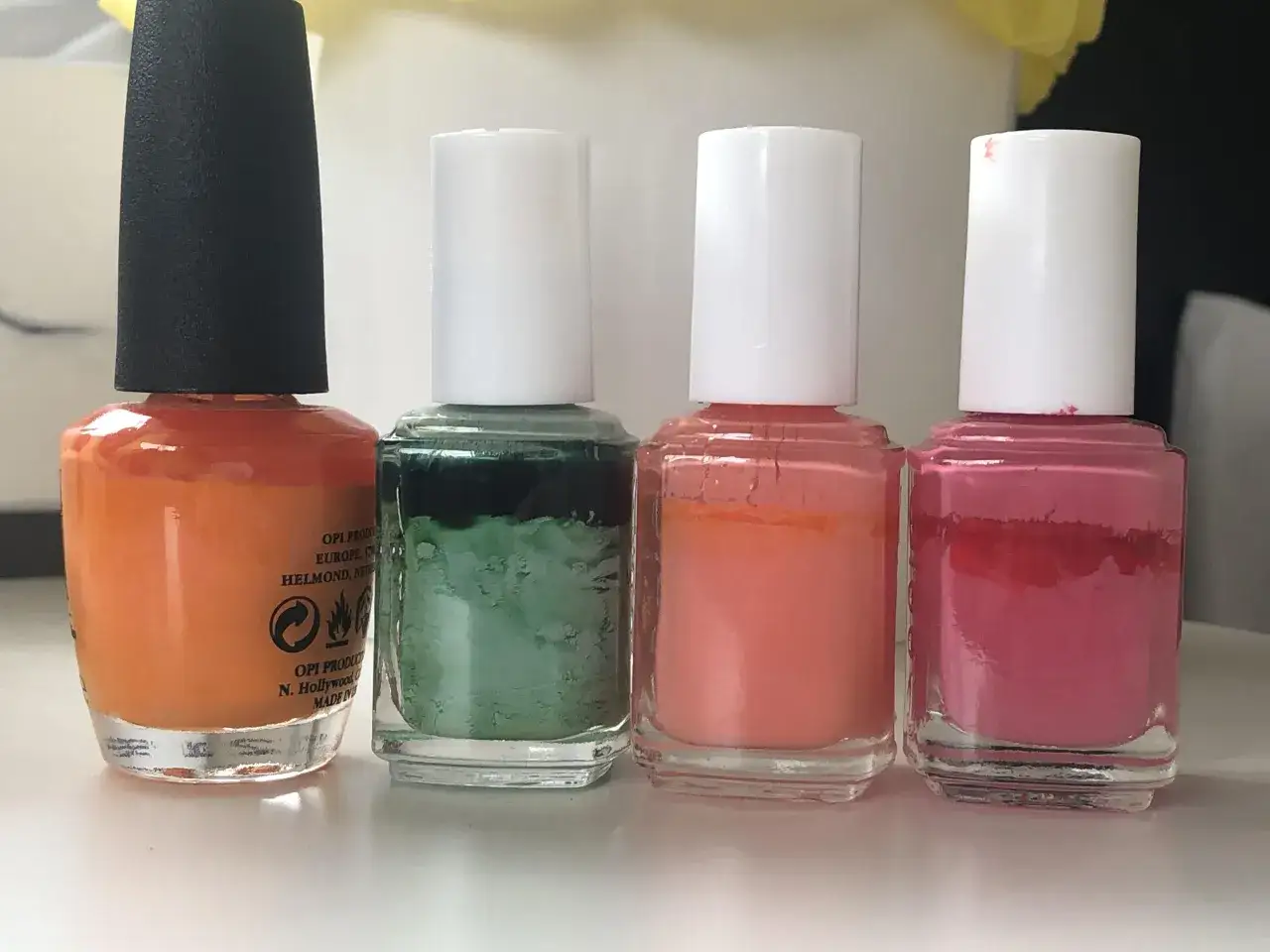 Vier Nagellackflaschen in Orange, Gr&uuml;n, Koralle und Pink. Man sieht, dass Nagellack ablaufen kann, da die Textur und Farbe sich ver&auml;ndert haben.