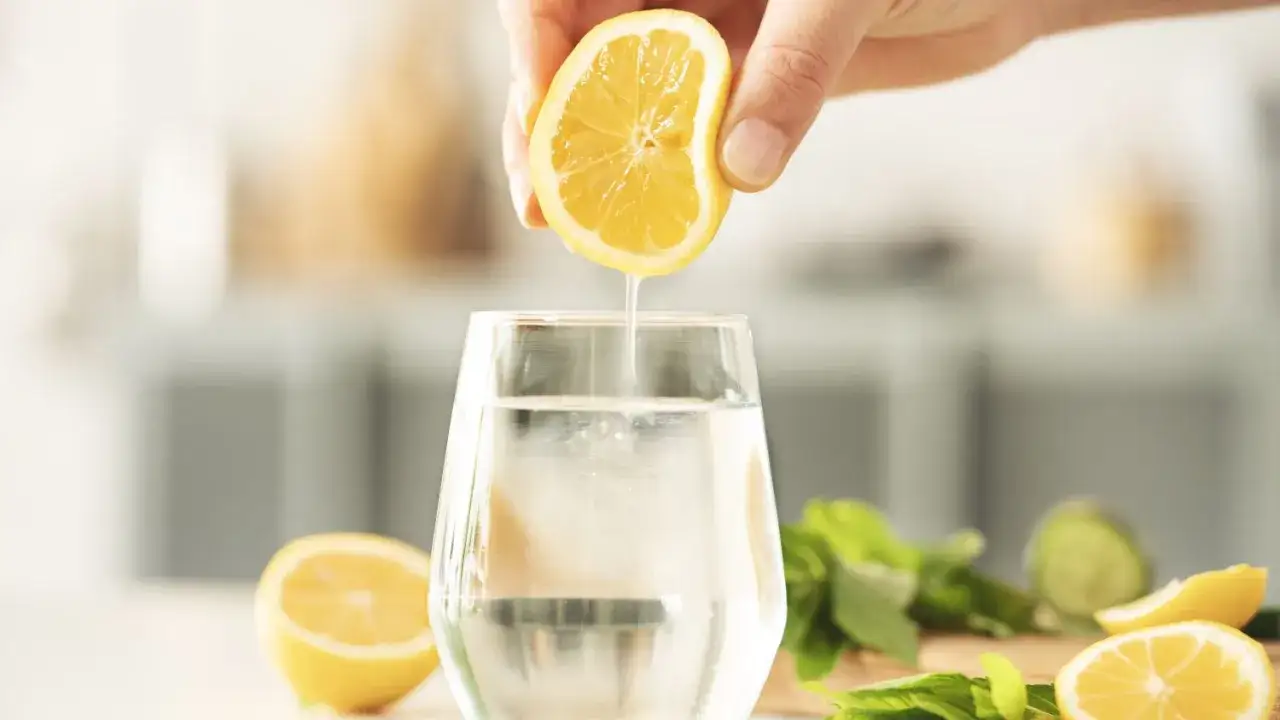 Eine Hand presst eine Zitronenhälfte aus, deren Saft in ein Glas Wasser tropft. Frisches Zitronenwasser morgens für einen guten Start.