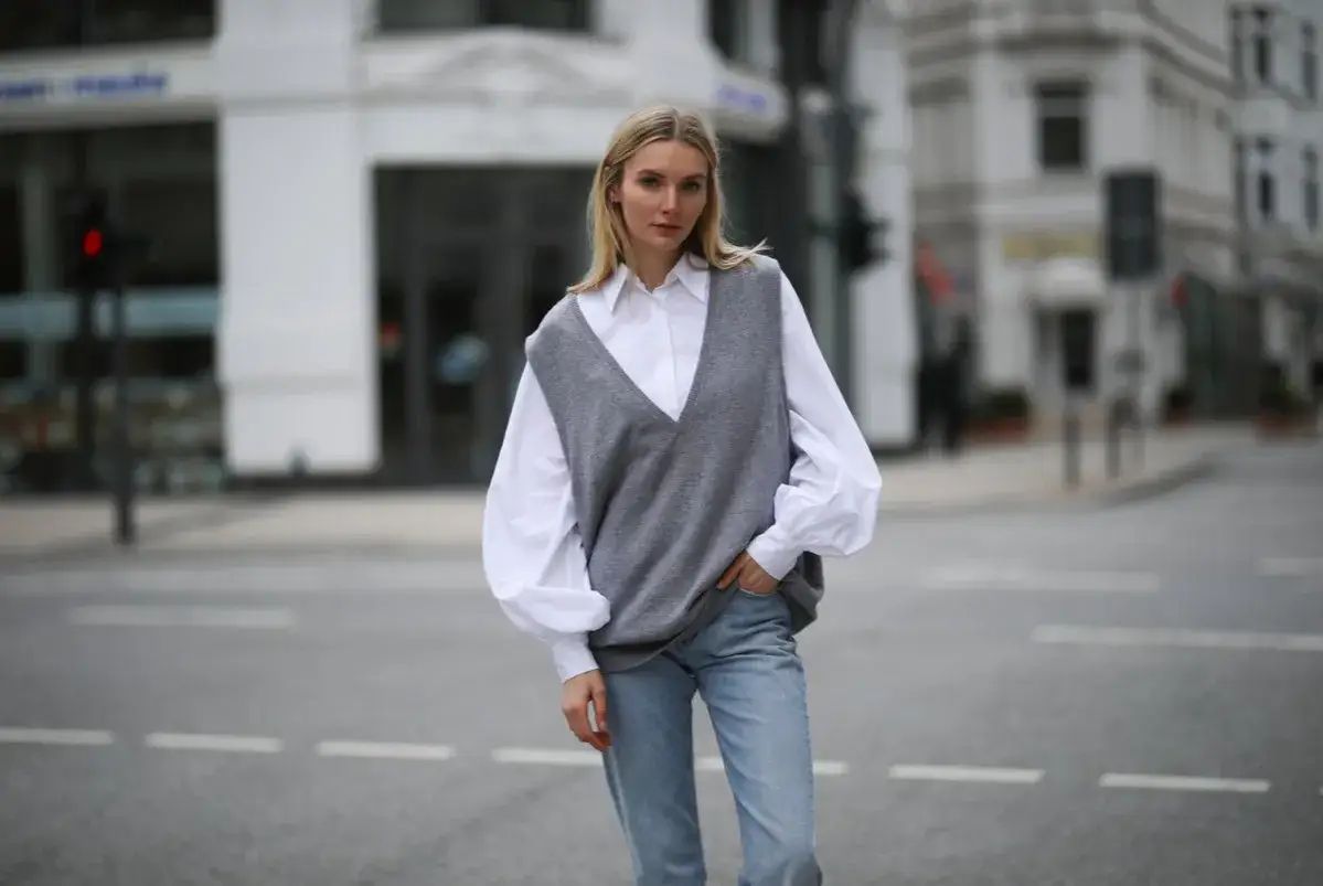 Frau in grauem V-Ausschnitt-Pullunder &uuml;ber wei&szlig;er Bluse und Jeans. Ein l&auml;ssiger Look, wie man einen Pullunder kombinieren kann.