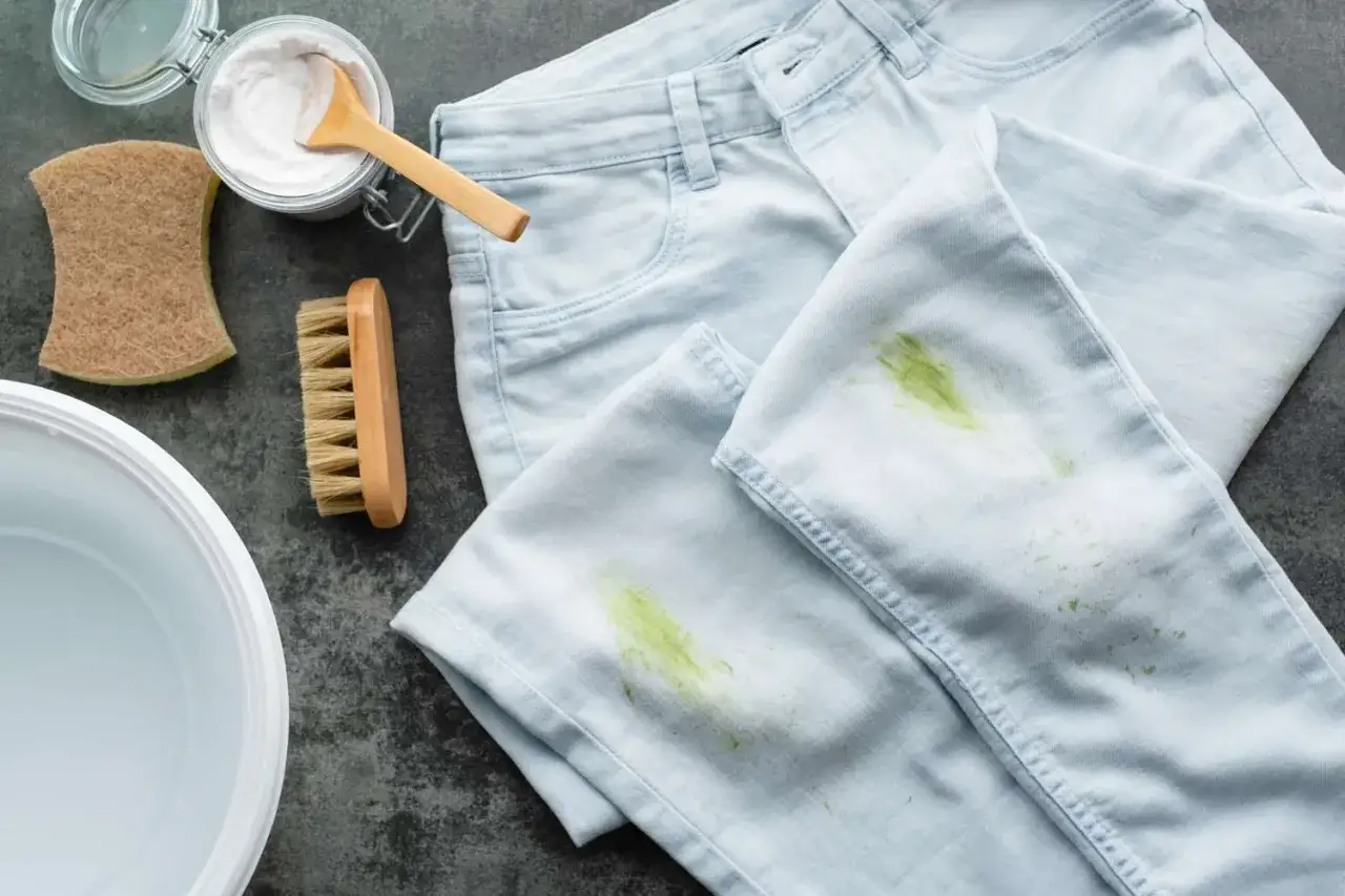 Hellblaue Jeans mit grünen Grasflecken, daneben eine Schüssel, Bürste, Schwamm und Natron.