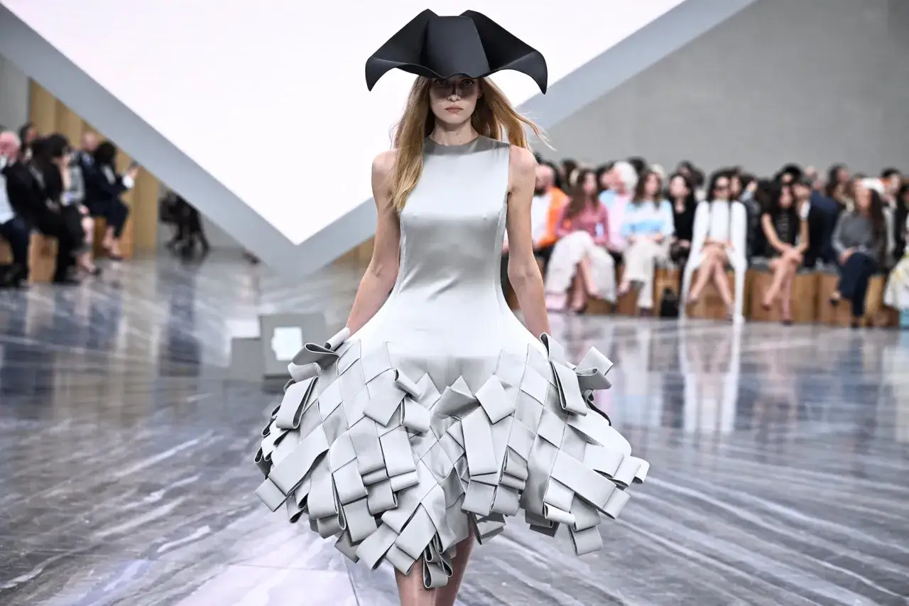Ein Model pr&auml;sentiert eine avantgardistische Kreation von Jonathan Anderson: ein schlichtes Kleid mit dramatischem, gefaltetem Rock und einem auff&auml;lligen Hut.
