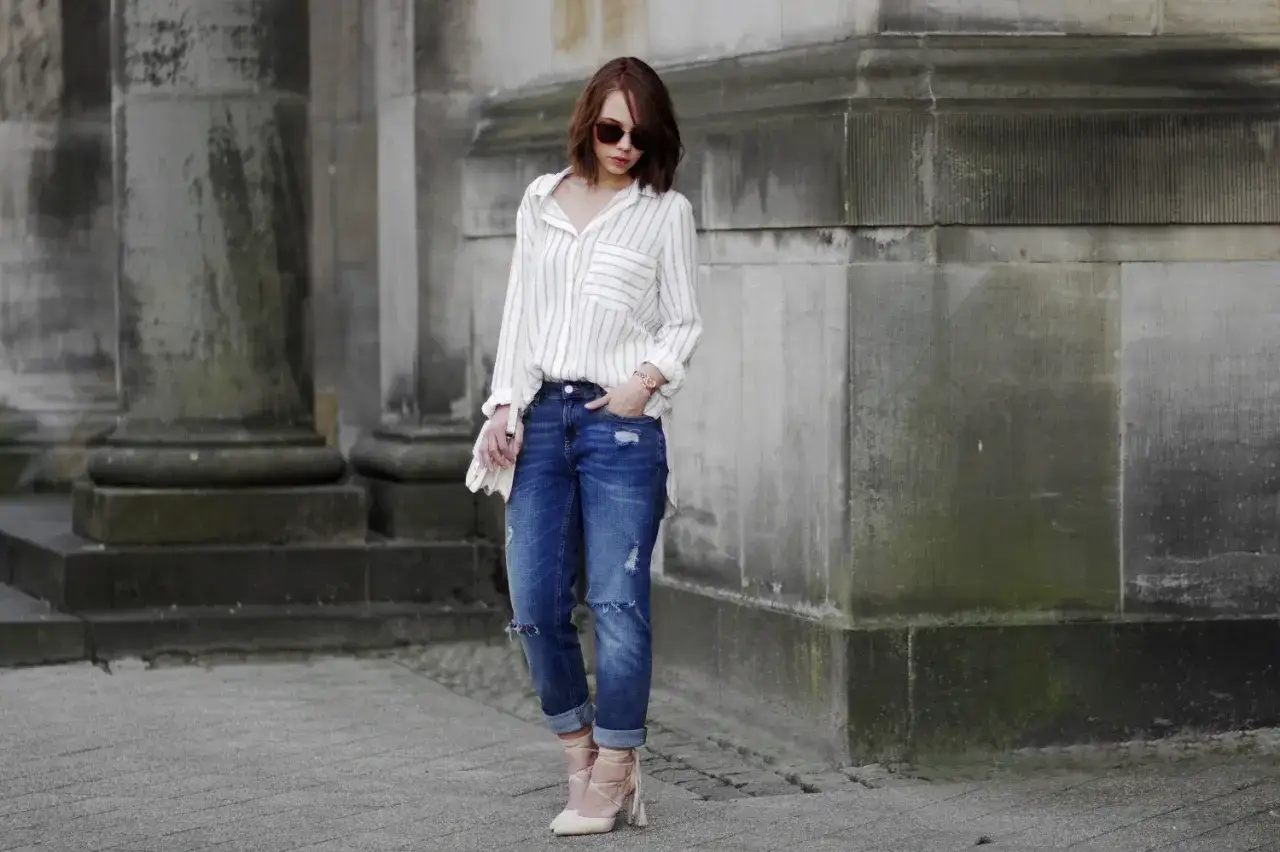 Streifenbluse mit zerrissener Jeans kombinieren: Ein l&auml;ssiger Look mit Absatzschuhen vor einer Steinfassade.